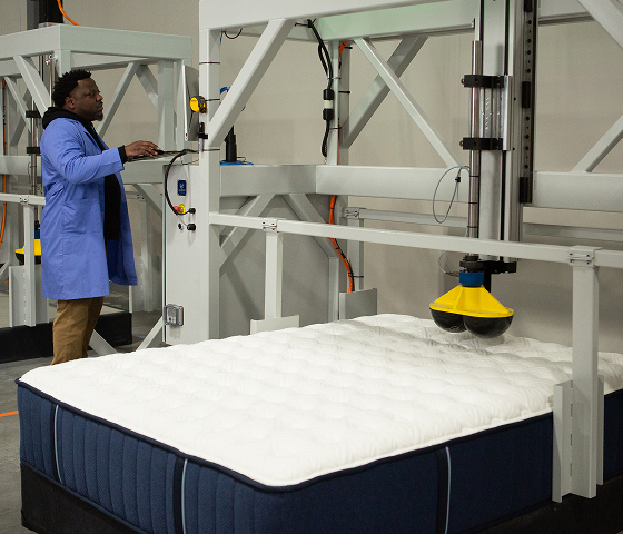 A man in a blue lab coat operates a large mattress-testing machine with a white mattress.
